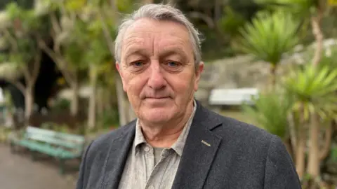 BBC Anthony Slaughter, standing in a public park in Penarth, in the Vale of Glamorgan, looking at the camera. There are palm trees and a park bench in the background. He is wearing a light grey shirt, open at the neck, and a dark grey jacket. He has short, grey hair.