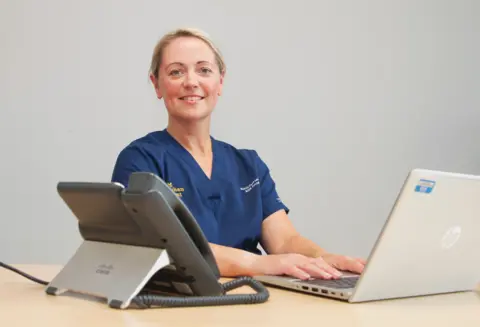 Dr Catherine Monaghan has blonde hair and is smiling. She is wearing a blue nurse's uniform and is sitting at a desk with a laptop and a large phone.