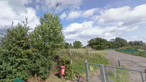 Google The entrance to Hook Road Arena, which is open space for the community between Chessington Road and Hook Road in Epsom. A metal gate is next to trees and plants. Trees and green grass in the background.