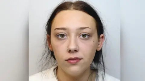 Wiltshire Police Custody image of Drew Stanton-Foreman. She is wearing a white jumper and has dark hair tied back and ticked behind her ears. She has one piercing under her right eye, and a nose ring. 