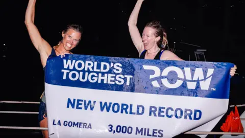 World's Toughest Row The picture shows two triumphant rowers standing side by side at night, both raising an arm in celebration while holding a large banner across the front of their bodies. The banner is wet, as if freshly splashed by sea water, and reads World’s Toughest Row and New World Record with the route labelled La Gomera 3,000 miles.
