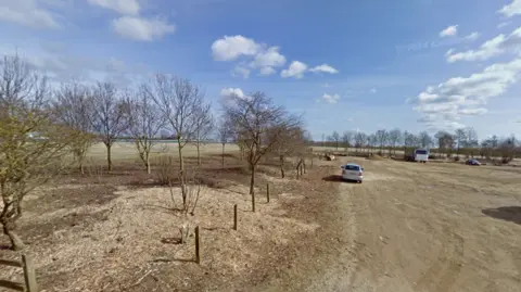 Image of the site, there is a car park to the right and some trees and grass to the left.