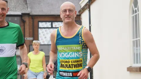 Colin Bailey A man running outside while wearing a running vest. The man has a bald head and glasses. He is wearing a turquoise vest that says "Samaritans", "Noggins", and "Custard Corner Marathon" on it in capital letters.