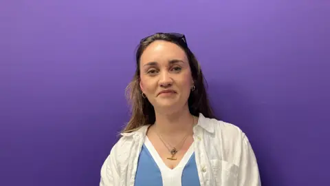 BBC Lara Vafiadis pictured in front of a purple background. She has long brown hair which is held back by a pair of sunglasses. She is wearing a gold necklace that sits above her light blue and white top. She is wearing a white linen shirt over the top.