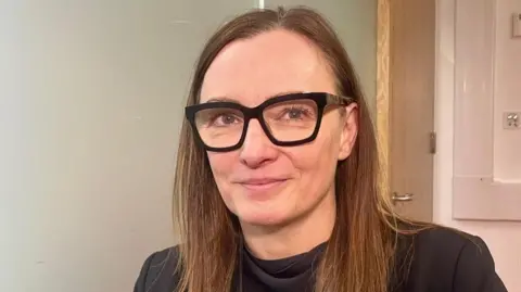 Sineidin Corrins in a room with plain beige walls and a door behind her. She wears dark glasses and a black top