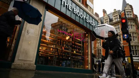 Woman walks with umbrella in front of a Body Shop store on a rainy day