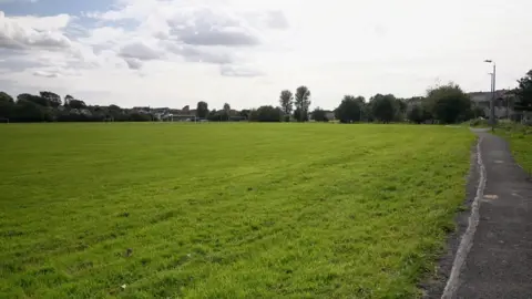 Playing fields made up of mostly grass, with a path on the right and trees, buildings and football posts in the distance