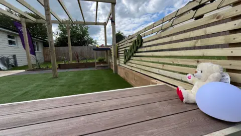 A photograph of the astroturf garden with a teddy bear in the foreground on a wooden bench. A wooden fence is on the right of the picture with Lucas's name in astroturf attached to the fence. 