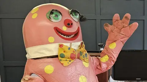 A large, pink costume is pictured with big googly eyes, a yellow bow tie and dots. It is raising his arm up with his hands out, waving.