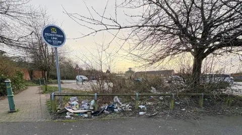 Abigail Spurrier Green grass leading to the car park of Morrisons supermarket in St Helens is filled with litter. The supermarket is seen in the background on a clear day.