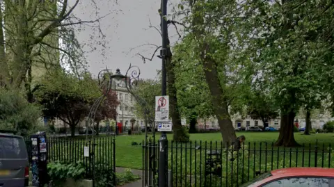 Portland Square, a small grassy park surrounded by black metal fencing. There are several trees in the park and cars parked outside it.
