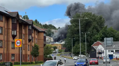 Dense black smoke pours into the sky from an apparent fire behind trees and low-rise buildings beside a curving road. Several cars are driving along the street, while brick apartment blocks line the left side of the image. The smoke contrasts sharply against patches of blue sky and white cloud overhead.