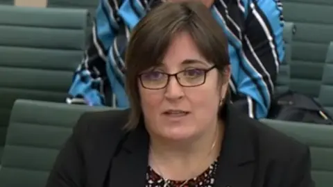 Catherine McLeod is sat appearing in front of MPs at the select committee. She is wearing a dark jacket, a gold necklace and dark glasses. 