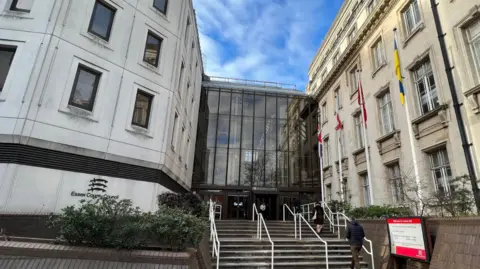 The entrance to County Hall in Chelmsford. It is a four or five-storey building. The facade is a mix of stone and glass. There are steps and white railings leading up to the entrance. There are flags on poles located outside.