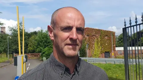 A man with a shaved head with light facial hair stares at the camera as he wears a black and white checked shirt. Behind his a building, black gates, and yellow barriers.