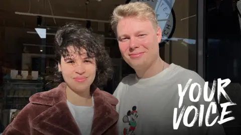 Emily Meekel and Victor Buitenhuis - she has dark hair and a maroon coat, he has blond hair and a white top with a picture of Mickey Mouse playing tennis on it. They are standing outside a bookshop/café. 
