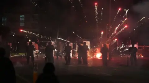 PA A street at night-time. The backs of silhouetted riot police, holding transparent riot shields, can be seen from behind. In front of them, a firework has just exploded, sending a number of red sparks shooting in all directions. In the foreground the silhouettes of a few onlookers can be seen.