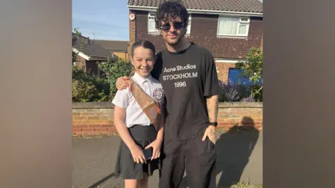 Contributed Tom Grennan wearing a brown T-shirt stands in a residential street with houses behind him. He has an arm round Orla who is in her school uniform. Orla also has a birthday sash around her white shirt and wears a badge saying 11th birthday.