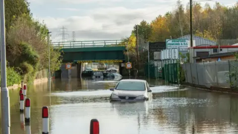 A car is submerged in flood water. In the distance other cars appear to be stuck in deep water under a bridge.