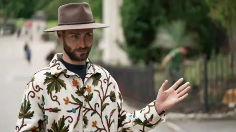 A man wearing a wide-brim brown hat and plant patterned shirt gestures with one hand.
