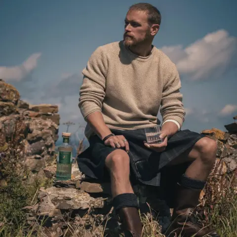 The Sassenach Sam Heughan wearing a kilt, sitting on a low wall holding a glass of gin.
