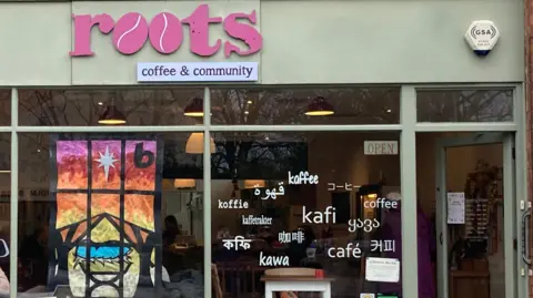 The front of a cafe - "Roots" is in pink writing with "coffee & community" underneath. Below you can see two windows, one with a colourful painted picture. The other window has the word "coffee" on it in multiple languages. The cafe is painted a light shade of green. 