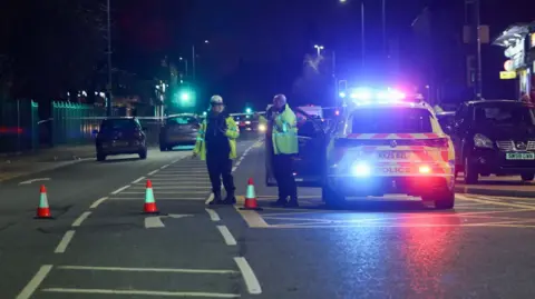 Two police officers stand by a cordon with bollards also sealing off the scene