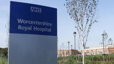 Tom Edwards A blue and grey sign outside a red building reads NHS Worcestershire Royal Hospital. In the background, there is road with a zebra crossing, some flower beds with bushes and trees, and a large brick building.