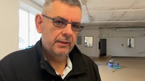 Peter Martin in a black jacket wearing glasses stood inside a community hall