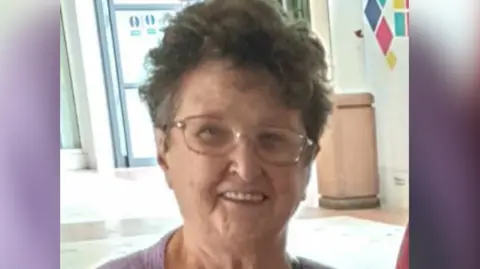 North Wales Police Celia Adams smiles at the camera. She wears glasses and has short dark grey hair, and is wearing a lilac cardigan