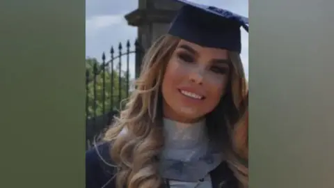 Family Handout Ashley Dale wearing a black university mortarboard and a grey top and black gown. She has long blonde hair and is smiling at the camera.