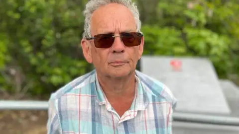 David Pracy standing in front of the war memorial. He has short white hair and is wearing dark glasses and an open-necked shirt in blue, grey, white and red plaid. 