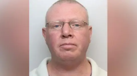 A mugshot of a man who has very short grey hair, flushed white skin, grey eyes and gold rimmed spectacles.