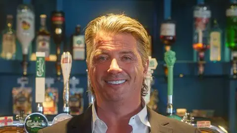 Indivisuals/Matt Filby A head and shoulders image of a man wearing a suit jacket over a white shirt while smiling. He is standing in front of a bar.