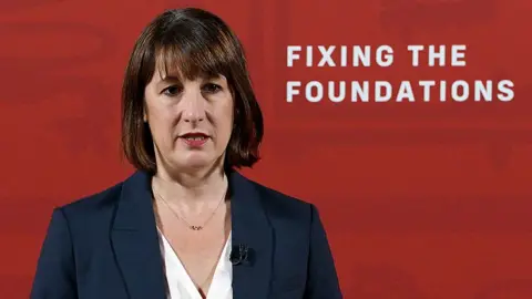 Getty Images Rachel Reeves stands in front of a red wall at a press conference