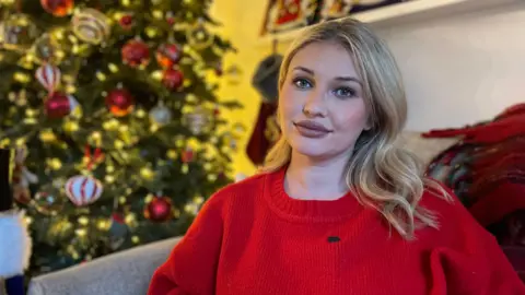 Amy Hart sitting in her living room. She has a red wooly jumper and blonde hair looking at the camera with a serious expression. Behind her is a lit up Christmas tree with decorations on.