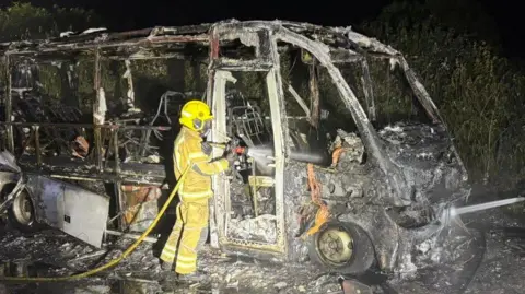 DWFRS Charred shell of bus with a firefighter standing by the driver's door.