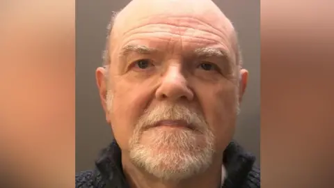 A mugshot of a elderly man, mostly bald and with a close cropped grey beard. The collar of a dark grey jumper or fleece can be seen. His expression is serious.