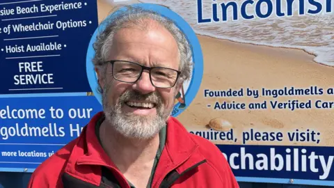 Lincolnshire County Council A head and shoulders shot of a man with grey hair and black rectangular glasses and a short beard and moustache smiling at the camera. He is wearing a red fleece. There is a large information board behind him.