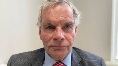 Sharon Edwards/BBC A close-up portrait of a man looking directly into the camera with a serious expression. He has short grey hair and is wearing a grey blazer and light blue shirt. The wall in the background is white.