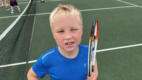 John Devine/BBC Eight-year-old Ruben Smith has short blonde hair and a blue and white t-shirt on, he is holding a tennis racquet under his left arm, and behind him is the tennis court.
