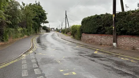 Luke Deal/BBC A road where there are yellow zig zag lines painted on the surface, but they are slightly faded. It is a grey day and it is raining. The road is lined by walls, buildings and electricity pylons.
