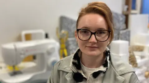 Jenny Kirk/BBC Kirstin Leigh. A woman with ginger, copper hair which is tied back. She is looking at the camera and is wearing a black and white fleece with a grey jacket. She is also wearing black glasses. She is sat in her studio and a sewing machine can be seen on a table behind her. 