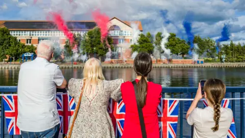 Stockton Council A middle-aged man and woman, a teenage girl and a younger girl are standing by the river as they watch and take photos of the colourful smoke display on the other side of the river bank. Union Jacks are hanging form the banister. 