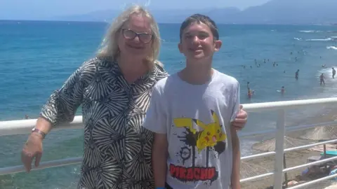 Teresa A woman poses for a photo with her arm around her son. Both are smiling, with a beach and swimmers in the background. The weather looks warm, with both wearing summer clothing.