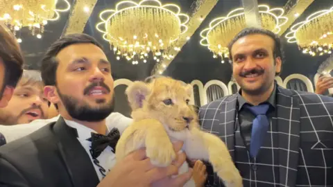 YouTube / Rajab's family rajab butt, in a suit, holding a lion baby. Omar doll wearing a suit is standing next to them and smiling