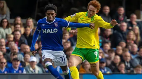 Getty Images An Ipswich Town football player in a blue shirt and white shorts during a tackle with a Norwich City player in a yellow and green kit. Fans sit in the stands behind them watching on.
