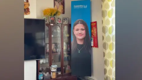 Jennifer Marlow A blue Coventry University poster with Jennifer Marlow's face on is in the corner of the living room. A cabinet with ornaments is to its left and further to the left is a black TV hanging on the wall.