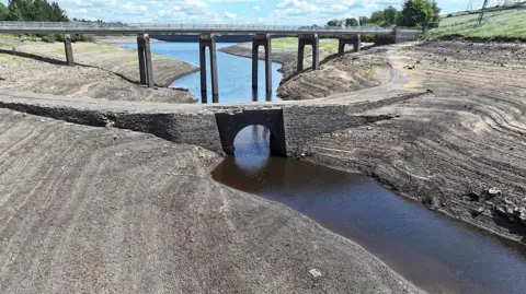 A dried-up bed of a reservoir with only a narrow stream of water flowing through the middle. In the background there is a bridge and a blue sky with scattered clouds.