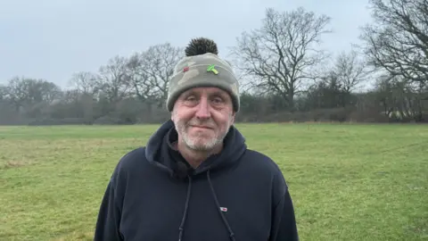 A man in a black hoodie and khaki beanie hat stood outside in a park, looking directly at the camera